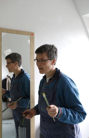 Karin Petereit in Arbeitskleidung an der Staffelei mit Pinsel vor Spiegel
